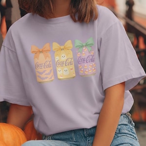 May include: A light purple t-shirt featuring three Coca-Cola cans with Halloween-themed designs. Each can has a bow and is decorated with bats, ghosts, and candy corn. The Coca-Cola logo is visible on each can.