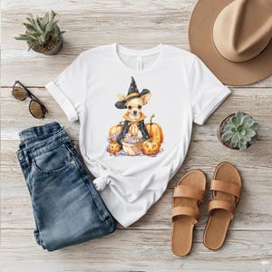 May include: White t-shirt featuring a watercolor illustration of a Chihuahua dressed as a witch, surrounded by pumpkins and flowers. The design is set against a wooden background, with jeans, sandals, and a hat.