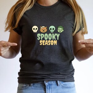Puede incluir: Camiseta gris oscuro con un diseño temático de Halloween. La camiseta muestra una calavera, un lobo con una venda, una calavera verde y una cara de zombi sobre las palabras "Spooky Season" en verde y naranja.