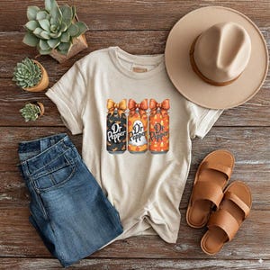 May include: Beige t-shirt featuring three Dr. Pepper cans with Halloween-themed designs. The cans have orange bows and are decorated with bats, ghosts, and candy corn. The shirt is paired with blue jeans, brown sandals, and a beige hat.
