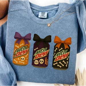 May include: A light blue sweatshirt featuring three Halloween-themed Mountain Dew can designs. Each can has a different color scheme, bow, and bone/skull accents. The text "MTN DEW" is prominently displayed on each can.