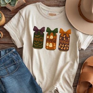 May include: Beige t-shirt featuring three Coca-Cola can designs with Halloween themes. The cans have purple, green, and orange bows. The designs include bats, ghosts, and candy corn. The shirt is paired with blue jeans, a hat, and sandals.