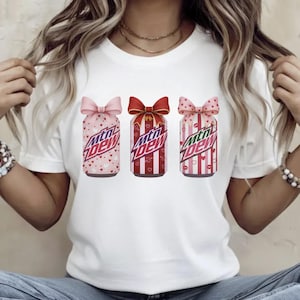 May include: White t-shirt featuring three stylized Mountain Dew cans with bow accents. The cans have heart and stripe patterns in pink and red, with the Mountain Dew logo prominently displayed.