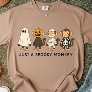 May include: Tan t-shirt featuring a graphic of four cartoon monkeys dressed as a ghost, pumpkin, mummy, and skeleton. The text "JUST A SPOOKY MONKEY" is printed below the monkeys. A silver necklace is visible at the neckline.