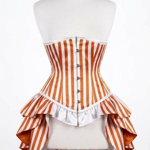 May include: A strapless corset featuring vertical orange and white stripes. The garment includes a front metal busk closure, a white ruffled trim along the waistline, and a high-low peplum hemline that extends into a long, flowing back panel.
