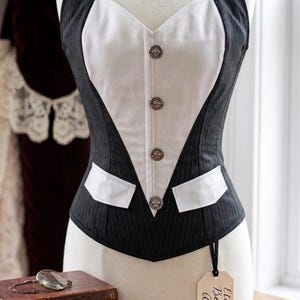 May include: A sleeveless corset-style vest featuring a black pinstriped fabric with a contrasting white V-shaped front panel. The garment includes four circular metallic buttons down the center and two white pocket flaps at the waistline.