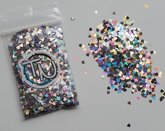 Pode incluir: Um saco de plástico transparente cheio de glitter em forma de coração iridescente. O glitter é uma mistura de prata, azul, verde e rosa. O saco tem um rótulo branco com o texto "TU" em um círculo azul.