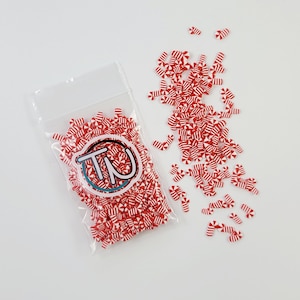 May include: A clear plastic bag filled with red and white candy cane sprinkles. The sprinkles are scattered around the bag on a white surface.