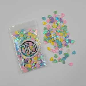 May include: A clear plastic bag filled with iridescent pastel-colored seashell-shaped glitter. The glitter is scattered on a white surface.