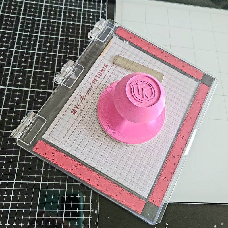 May include: A clear acrylic stamping block with a pink rubber stamp featuring the text "MY Sweet PETUNIA" and a pink knob. The block has a grid pattern and a ruler on the edge.