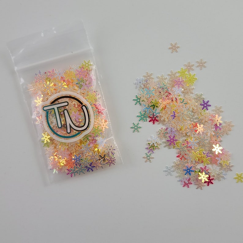 May include: A clear plastic bag filled with iridescent snowflake-shaped glitter. The glitter is scattered on a white surface. The snowflakes are in various pastel colors, including pink, yellow, and blue. The bag has a circular logo with the letters "TN".