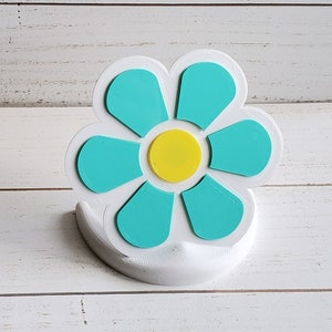 May include: A white 3D printed flower with a yellow center and teal petals. The flower is on a white base.