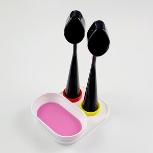 Op de afbeelding: Witte plasticen standaard met twee zwarte make-up kwasten. De standaard heeft een roze ovaalvormige schaal aan de voorkant.