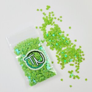 May include: A clear plastic bag filled with green glitter shaped like four-leaf clovers. Some of the glitter is scattered on a white surface.