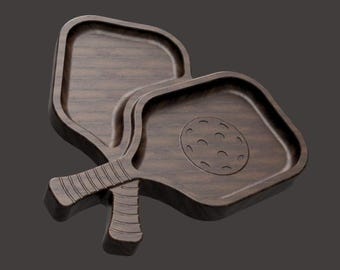 May include: Two wooden pickleball paddle-shaped trays. The trays are dark brown with a wood grain pattern. One tray has a carved pickleball design. The handles are textured for grip.