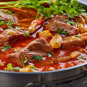 May include: A close-up of a pot of red stew with chunks of meat and green vegetables. The stew is topped with fresh parsley.