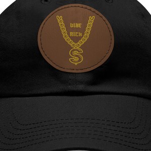 May include: Black baseball cap with a brown patch featuring a gold chain and dollar sign design. The text "Vibe Rich" is above the chain. The cap has a curved brim and an adjustable strap.