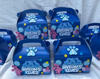 May include: Navy blue party favor boxes with handles, decorated with paw prints, the number 1, and the text "Aveon's Clues". Pink cartoon characters and blue and yellow circles accent the boxes. Party supplies.