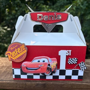 Puede incluir: Una caja de regalo blanca y roja con un diseño de Rayo McQueen. La caja tiene el nombre "Denis" en la parte superior, el número "1st" y banderas a cuadros. También es visible una pegatina de "Rust-eze".