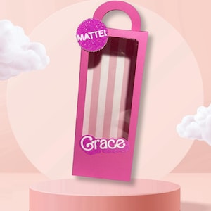 Puede incluir: Una caja rosa con asa, con una ventana transparente que revela un patrón de rayas. La caja tiene el nombre "Grace" y la palabra "Mattel". La caja se muestra sobre una plataforma rosa.