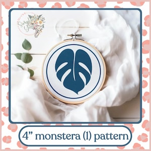 Puede incluir: Un aro de bordado de 10 cm con un fondo de tela blanca y un diseño de hoja de monstera azul oscuro. El aro está sobre una superficie de tela blanca.