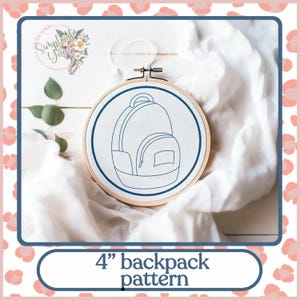 May include: A 4-inch embroidery hoop with a white fabric background. The fabric has a blue outline of a backpack with a zipper and a pocket.