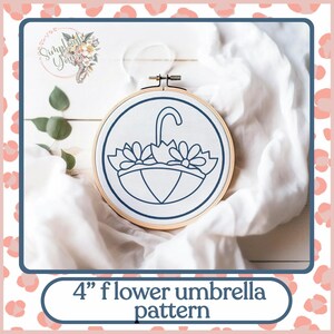 May include: A 4 inch embroidery hoop with a white fabric background. The fabric has a blue outline of a flower umbrella with three flowers on top.