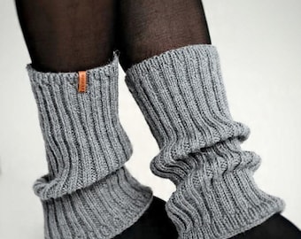 May include: A pair of grey ribbed knit leg warmers worn over black tights. The leg warmers feature a vertical ribbed texture and a small, rectangular brown leather-like brand tag attached to the upper edge of the left leg warmer.