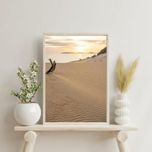 May include: A framed photograph of a sandy beach with a piece of driftwood in the foreground. The beach is empty and the sun is setting over the ocean in the background.