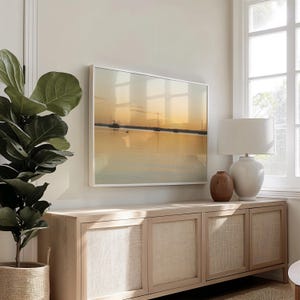 May include: A framed landscape art print depicting a serene sunset over a body of water, with silhouettes of boats. The artwork is displayed above a light wood cabinet with woven door panels, next to a potted plant and a white lamp.