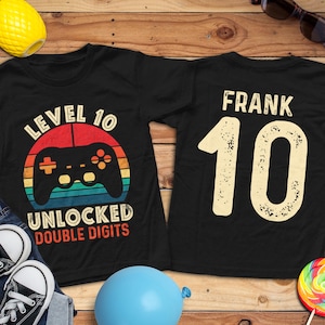 May include: Black t-shirts with a gaming theme. One shirt features a retro-style graphic with a controller and the text "LEVEL 10 UNLOCKED DOUBLE DIGITS." The other shirt has the name "FRANK" and the number "10."