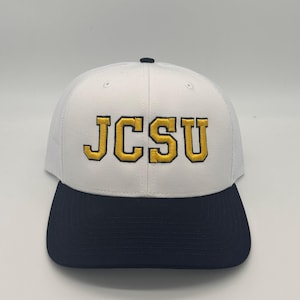 Può includere: Un cappellino da baseball bianco con visiera blu scuro e pannelli posteriori in rete. La parte anteriore presenta le lettere JCSU ricamate in giallo con un bordo blu scuro. Il cappellino ha un piccolo bottone blu scuro sulla sommità.