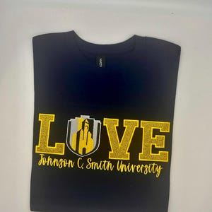 Op de afbeelding: Een marineblauw t-shirt met het woord LOVE in gouden glitterletters, waarbij de letter O is vervangen door een grijs en goud schildembleem met een gebouw. Onder de afbeelding staat de tekst Johnson C. Smith University in het goud.