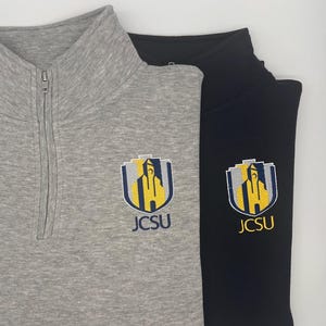 Peut inclure: Un pull à quart de zip gris chiné et un pull à quart de zip noir, tous deux dotés d'un col montant avec une fermeture éclair argentée. Chaque vêtement présente un logo écusson brodé en jaune, bleu et gris avec le texte JCSU en dessous.
