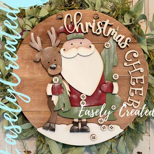 Può includere: Cartello natalizio in legno con Babbo Natale e una renna, con le parole "Christmas Cheer". Il cartello è circondato da verde e riporta il testo "Easely Created" e "Digital Cut File".