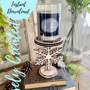 May include: A wooden snowflake candle holder with a white base and a brown snowflake design. A glass candle jar with a black label that says "Cozy Nights" is sitting on the holder. The candle holder is on top of a stack of books.