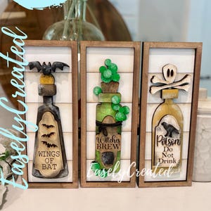 May include: Three wooden framed Halloween-themed signs. Each sign features a bottle design with decorative elements: a bat, cactus, and skull. The signs include the text "Wings of Bat", "Witches Brew", and "Poison Do Not Drink". The signs are labeled "Easely Created".
