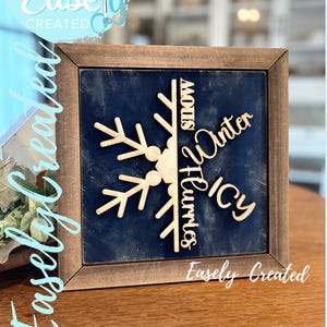 Peut inclure: Panneau encadré en bois avec un fond bleu marine et un motif de flocon de neige découpé au laser. Le flocon de neige porte les mots "Snow, Winter, Icy, Flurries" et le texte "Easely Created".