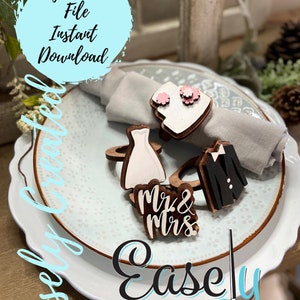 May include: Wooden napkin rings with a bride and groom design. The rings are painted white and black and have pink flowers. The rings are on a white plate with a gray napkin. The text "Mr. & Mrs." is on one of the rings. The text "Easley Created" is on the plate.
