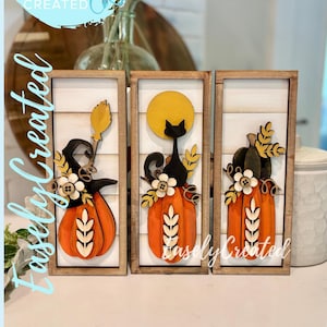 May include: Three wooden framed fall-themed wall art pieces. Each features an orange pumpkin with decorative elements like a witch's hat, black cat, and floral accents. The background is white with wooden slats. Text on the image reads "Digital Cut File Easely Created".