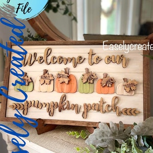 Peut inclure: Une pancarte en bois avec le texte "Bienvenue dans notre champ de citrouilles" et cinq citrouilles avec des noms dessus. La pancarte est sur un chevalet en bois.