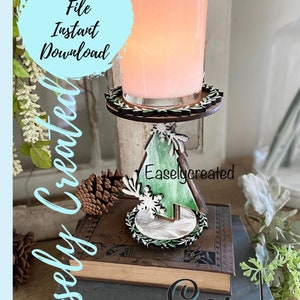 May include: A wooden candle holder shaped like a Christmas tree with a green and white painted design. The holder has a round base with a wreath design and a glass candle jar on top. The candle holder is sitting on a stack of books with the text "Easley Created" on the bottom book.