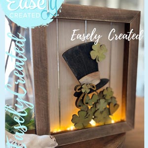 May include: Wooden framed St. Patrick's Day decoration featuring a black top hat with a green shamrock and a cluster of green shamrocks. The words "Easely Created" and "Digital Cut File" are visible.