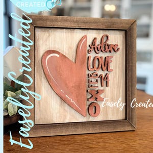 Op de afbeelding: Houten bord met een hartontwerp en de woorden "Adore Love 14 Feb XO". Het bord heeft een lichtbruine achtergrond, een donkerbruine lijst en de tekst "Easely Created". Geschikt voor Valentijnsdag.
