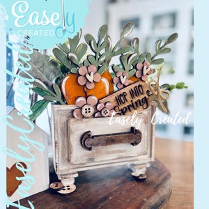 Peut inclure: Un tiroir décoratif en bois sur le thème du printemps. Il présente des carottes oranges, des fleurs roses et des feuilles vertes. Le tiroir a une poignée en métal et les mots "Hop Into Spring" sur une étiquette. Le texte "Easely Created" est également visible.