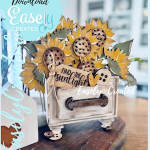 May include: A wooden decorative arrangement featuring sunflowers, leaves, and a small drawer. The sunflowers have yellow petals and brown centers. The drawer has a distressed white finish and the words "Find the Sunlight" are on a wooden tag.