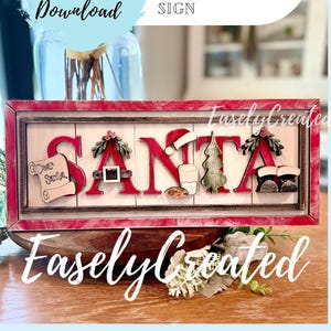May include: A rectangular wooden sign with a red frame featuring the word SANTA in large red letters. Each letter is decorated with holiday icons including a scroll, a belt buckle, a glass of milk, a cookie, a pine tree, and boots.