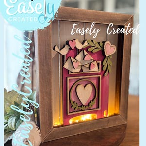 Puede incluir: Caja de sombra con marco de madera con interior rosa, con pájaros, corazones y follaje de madera cortados con láser. El texto "Easely Created" es visible en el marco y en un diseño circular. La caja está iluminada con luces cálidas.