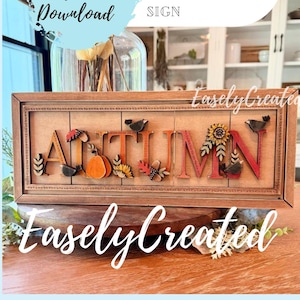May include: A rectangular wooden sign featuring the word AUTUMN in raised, multi-colored letters. Each letter is decorated with seasonal motifs including a pumpkin, sunflower, and birds. The sign has a dark wood frame with a stitched-look border.