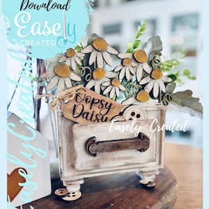 Op de afbeelding: Een rustieke houten lade met madeliefjes uitsparingen en een label met de tekst "Oopsy Daisy". De lade is wit met een metalen handvat en staat op een houten voet. De afbeelding bevat de tekst "Instant Digital Download Easely Created."